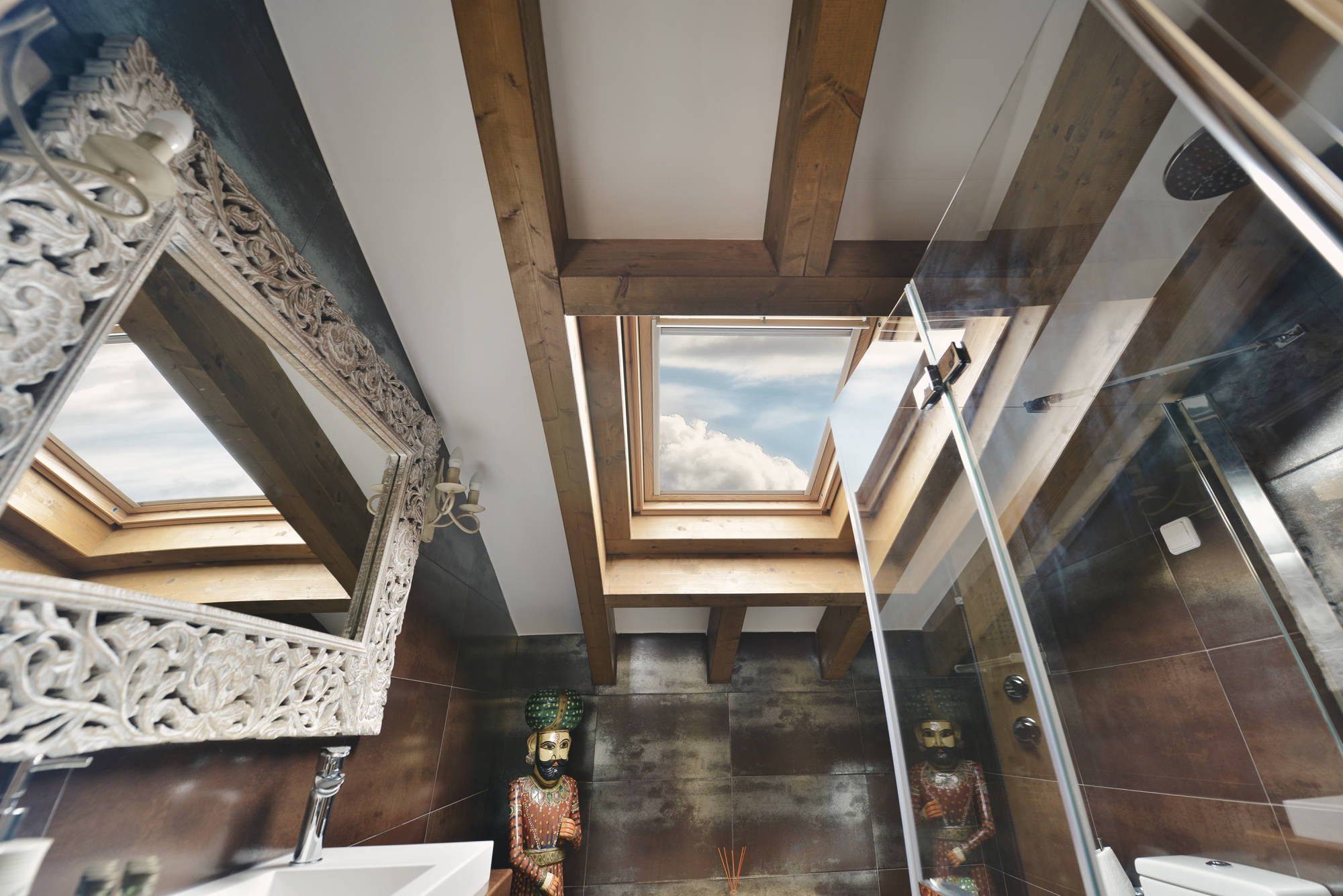 Cuarto de baño abuhardillado con tragaluz en el techo de vigas de madera, paredes de acabado oscuro, espejo tallado de estilo decorativo y elementos ornamentales exóticos junto a la ducha acristalada.