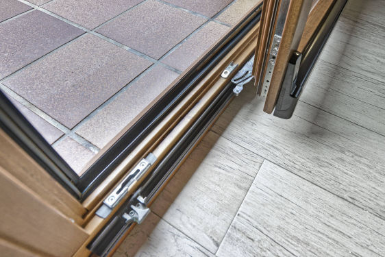 Detalle inferior de cerramiento corredero con perfilería de madera y herrajes metálicos visibles, sobre suelo interior cerámico y exterior en terraza.